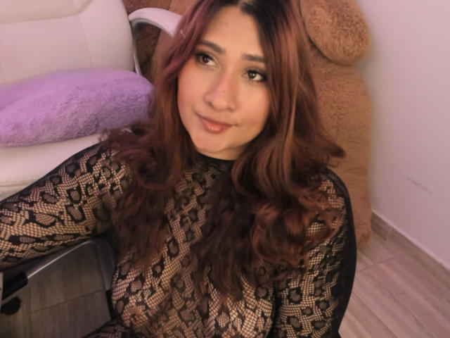 ElyRoss mit welligem, braunem Haar, trägt eine schwarze Spitzenbluse, die ihre Brüste zeigt, lächelt in die Kamera und sitzt auf einem Stuhl mit einem violetten Kissen und einem braunen Teddybär..