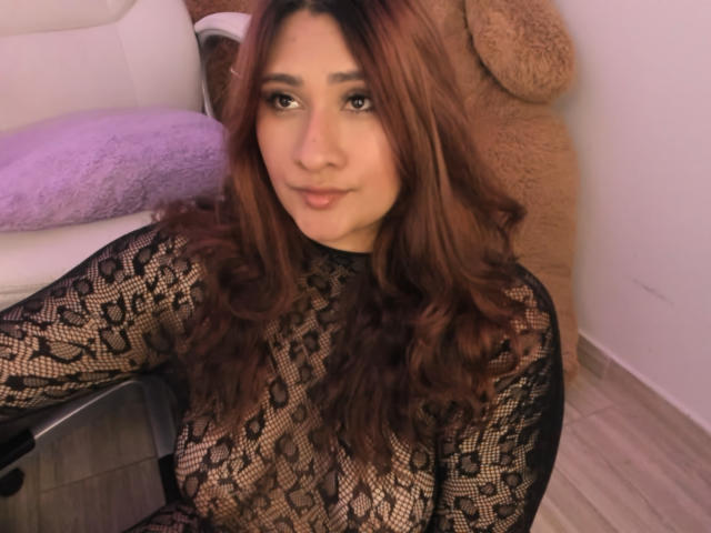 ElyRoss mit braunen, gewellten Haaren, trägt eine schwarze Spitzenbluse, die ihre Brüste zeigt, sitzt auf einem Stuhl, im Hintergrund ist ein flauschiger, brauner Teddybär zu sehen..
