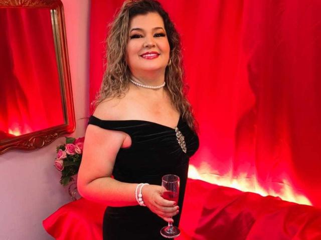 AdrianaPrettie with curly blonde hair, red lipstick, black off-shoulder dress, pearl necklace, bracelet, holding a champagne glass, smiling, red curtains, gold-framed mirror, pink roses.