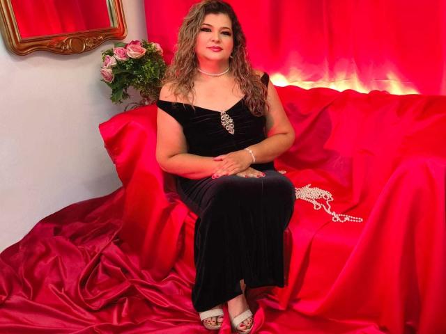 AdrianaPrettie with wavy brown hair, red lipstick, black off-shoulder dress, pearl necklace, white heels, sitting on red satin-covered couch, bouquet of pink roses on left.