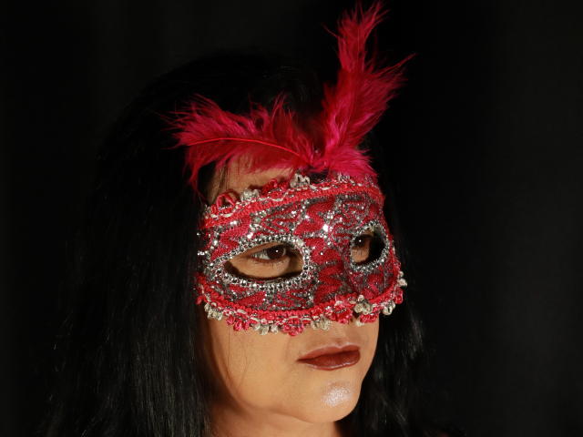 VonneLucia, con cabello negro oscuro, lleva una máscara de carnaval roja, adornada con plumas rosas y con brillantes, y tiene pintalabios rojo oscuro, mirando de lado..