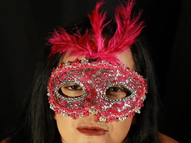 O rosto de VonneLucia, adornado com uma máscara de carnaval vermelha e brilhante, com penas rosa e pedras preciosas cintilantes, cabelo preto e batom marrom..