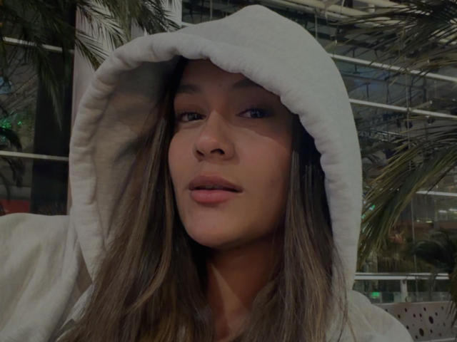 ThaliaAres with brown hair, wearing a white hoodie with the hood up, has a slight smile, and a cute face, in a soft-lit, urban setting.