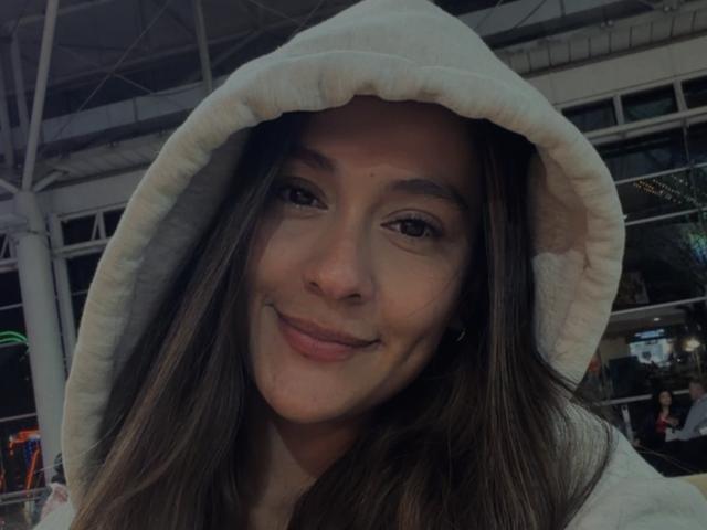 ThaliaAres with long brown hair, smiling, wearing a white hooded sweatshirt, has cute brown eyes and a small nose. Her ass is not visible.