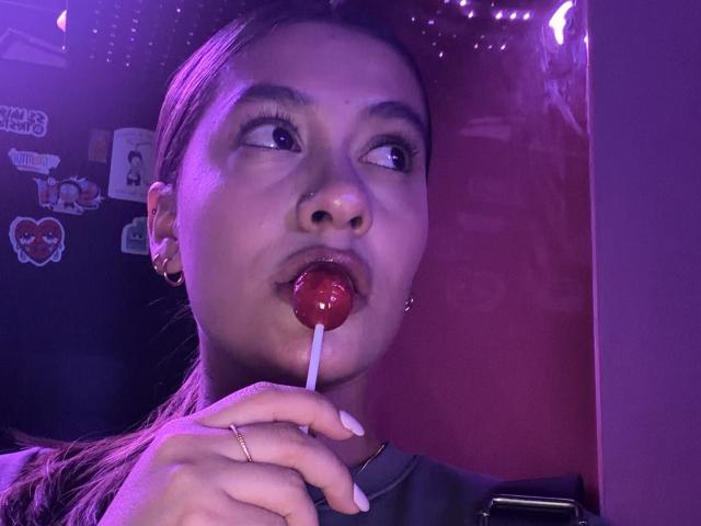 ThaliaAres with her lips around a red lollipop, holding it with her right hand, her gaze looking upward. Purple lighting casts a soft glow on her face.
