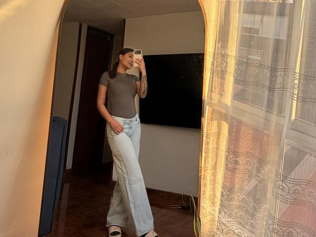 ThaliaAres with tattoos, wearing a gray tee, blue flared jeans, and sandals, taking a mirror selfie, showing her ass, in a room with a TV and curtain.