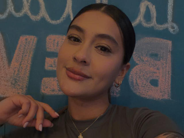 ThaliaAres with dark hair, brown eyes, and a small gold necklace, smiling with pink lips, wearing a gray shirt, and a hoop earring, resting her hand on her cheek.