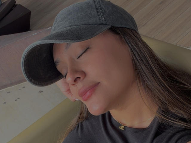 ThaliaAres with closed eyes, wearing a gray cap, black shirt, and a small gold necklace, has long brown hair and a slight smile, showing a relaxed, pretty face.