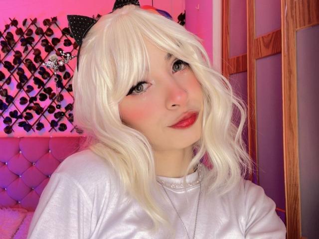 LaurenEvie69 with blonde wig, cat ears, red lipstick, and white shirt, showing cute face and pink blush, sitting in pink and purple lit room.