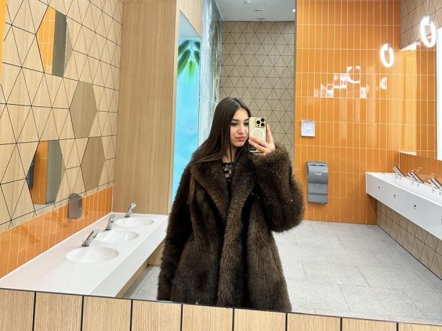 ShawneeLinderholm in a brown furry coat, taking a mirror selfie in a modern bathroom, holding a gold iPhone, with long dark hair, looking at the camera.
