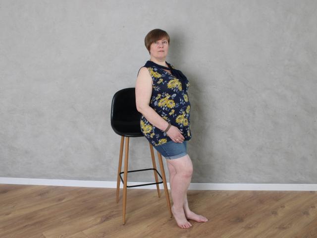 RadaChudna, middle-aged woman with short brown hair, standing barefoot, wearing a floral blue top and denim shorts, leaning on a black bar stool against a gray wall.