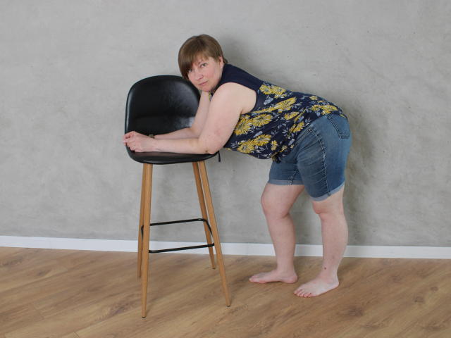 RadaChudna bending over a black bar stool, wearing a floral sleeveless top, blue denim shorts, barefoot on wooden floor, grey wall behind, her ass slightly visible.