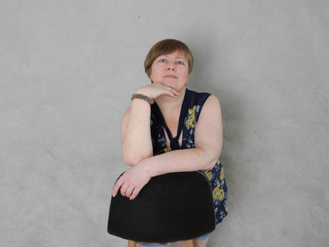 RadaChudna with short brown hair, blue floral sleeveless top, black purse, left arm resting on chest, right hand on purse, gray wall background, looking slightly up, bracelet on wrist.