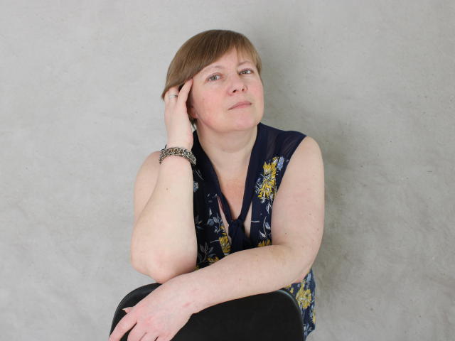 RadaChudna has short brown hair, fair skin, and is wearing a sleeveless navy floral top, black pants, and a silver bracelet, with a thoughtful expression, hand on cheek.