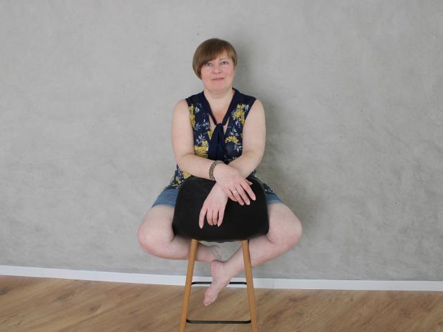 RadaChudna, chubby with short brown hair, sits on a wooden stool, legs spread, wearing a floral sleeveless top and denim shorts, black pillow on her ass, bare feet.