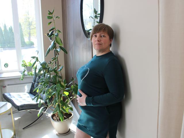 RadaChudna stands against a wall in a green long-sleeve dress, her right hand on her belly, with a plant to the left and a window behind her.