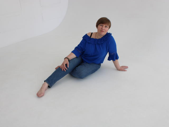 RadaChudna is sitting barefoot on a white floor, wearing a blue off-shoulder top and jeans, with short brown hair, and a bracelet on her right wrist.