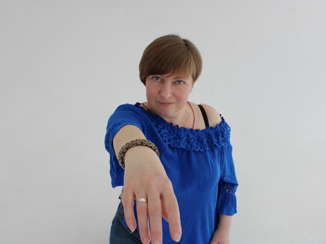 RadaChudna, con cabello castaño corto, una blusa azul con un hombro descubierto, extendiendo la mano derecha hacia adelante, llevando una pulsera y un anillo, sonriendo ligeramente, fondo blanco..