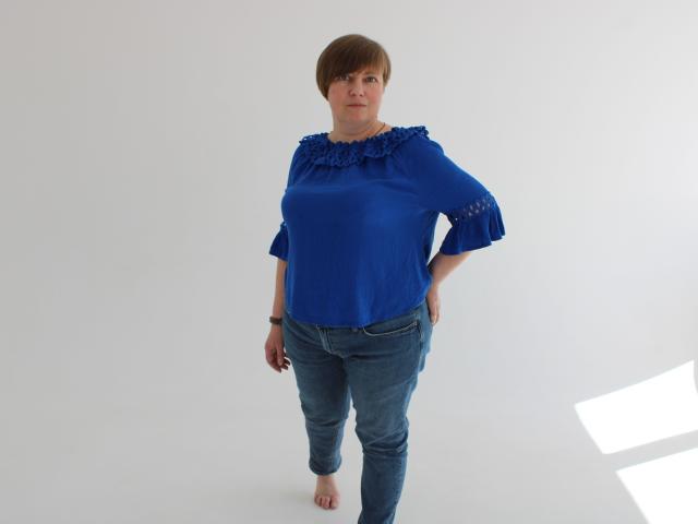 RadaChudna está de pie, descalza, vistiendo una blusa azul con encaje y jeans azules, con el pelo corto y castaño, con las manos en las caderas, mirando hacia adelante. Es una mujer de mediana edad, de figura curvilínea..