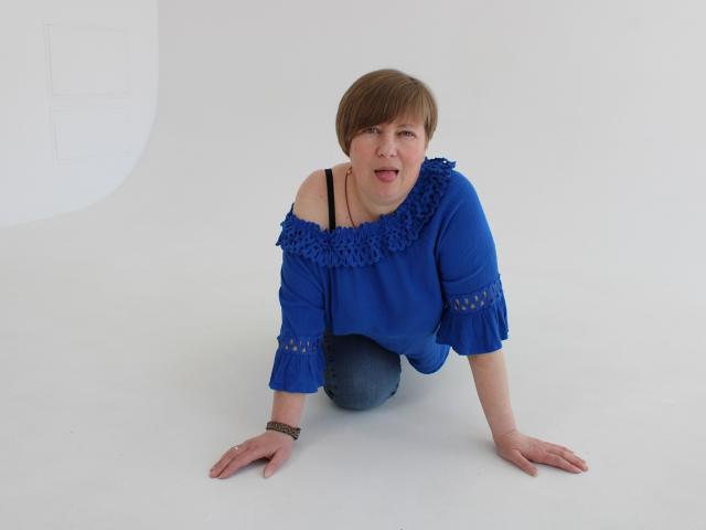 RadaChudna, a short-haired woman, kneels on white floor, wearing blue off-shoulder top with lace, black strap, and blue jeans, looking forward with mouth open.