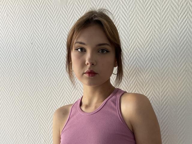 VenettaBershadsky with light brown hair, wearing a pink tank top, has small tits, subtle makeup, and a neutral expression, standing against a white, textured wall.