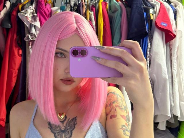 VenettaBershadsky with pink bob, holding purple phone, showing tattooed arm, butterfly chest tattoo, wearing blue top, in mirror selfie, showing pale skin, painted nails, heart necklace.