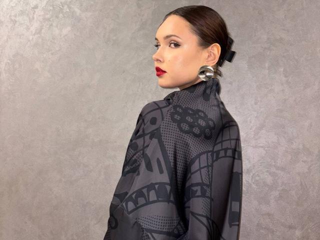 VenettaBershadsky with slicked-back brown hair, red lipstick, and black geometric-patterned shawl, wearing large metallic earrings, looking over shoulder.