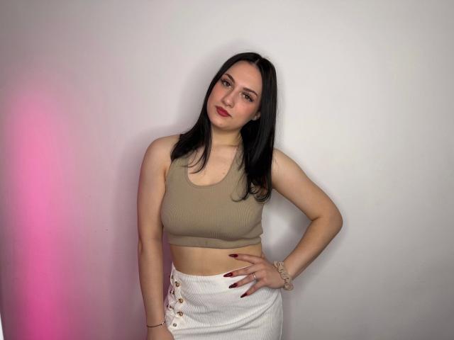 Darkling-hot with straight black hair, pale skin, and red lipstick, wearing a beige crop top and white skirt with button details, standing with hand on hip.