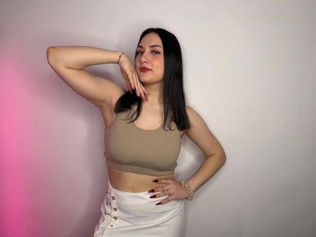 Darkling-hot with pale skin, black hair, red lipstick, wearing a beige sports bra and white button-up skirt, hand on hip, other hand touching chin, looking confident.