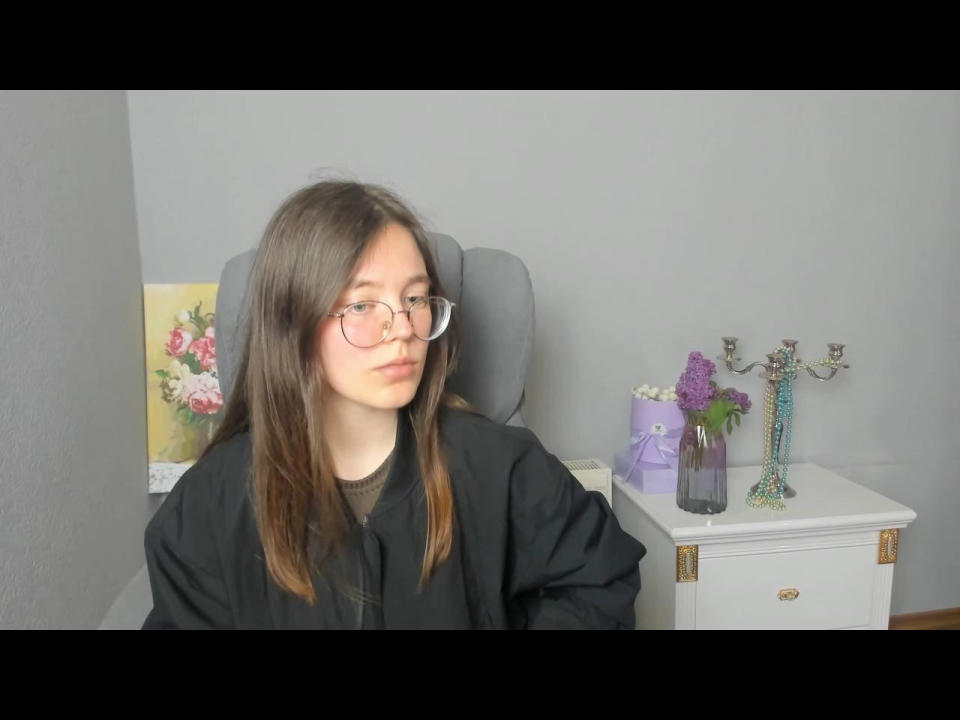 Raneji, com óculos e cabelo castanho, está sentada em uma cadeira, vestindo uma camisa preta, ao lado de uma mesa branca com um vaso roxo, um candelabro verde e flores lilás..