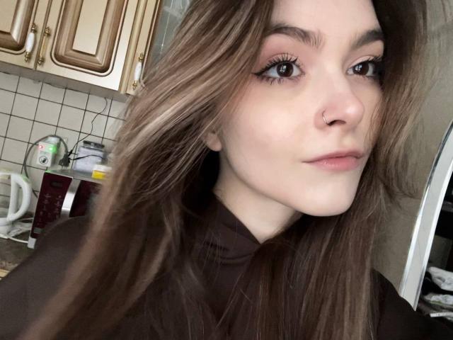 RonnaBarken has long brown hair, a nose ring, and wears black eyeliner. She's in a kitchen, looking at the camera with a slight, confident smile.