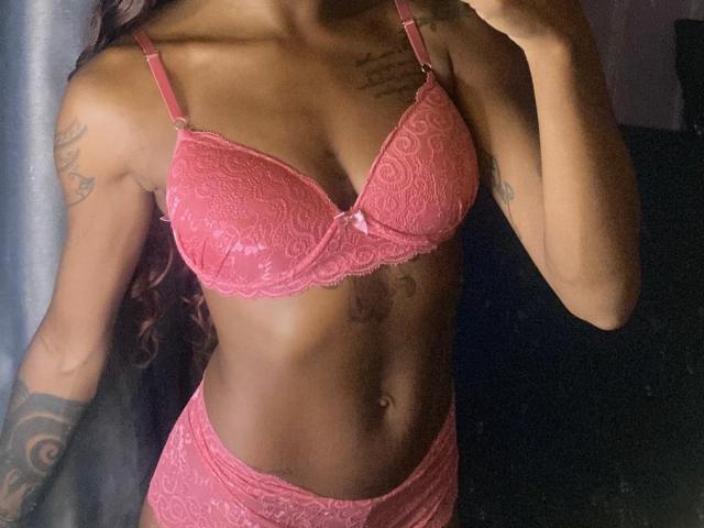 Savvana, vêtue de lingerie en dentelle rose, montre ses seins dans un soutien-gorge et une culotte assortis, avec des tatouages sur les bras et un corps mince, tonique et de couleur foncée..