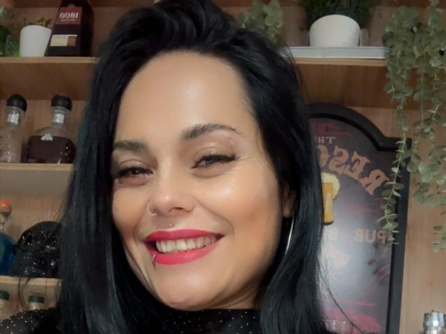 CharlotteCrofort, with black hair, red lipstick, nose ring, and smiling, showing a small scar above her right eyebrow. She's wearing a black top with small white dots.