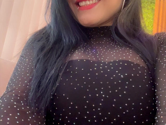 CharlotteCrofort with black speckled mesh top, showing tits, has long blue-black hair, smiling with red lipstick, visible teeth, and wearing large hoop earrings.