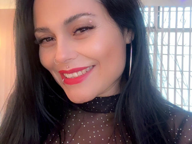 CharlotteCrofort has long black hair, red lipstick, a nose ring, and a smile, wearing a sheer black top with tiny white dots, and large hoop earrings.