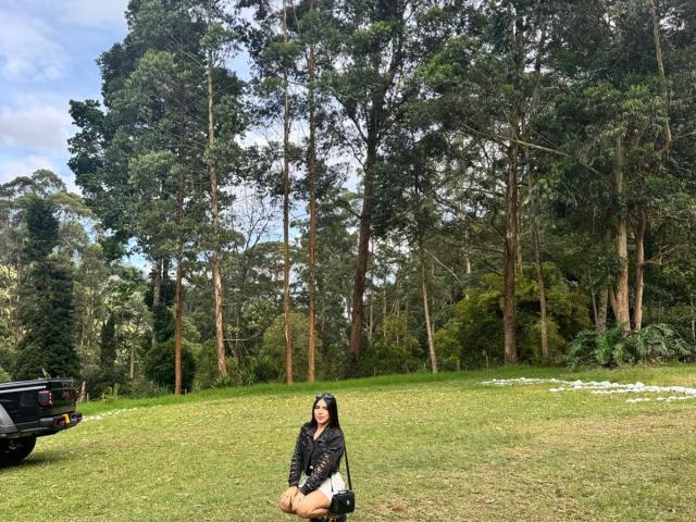SophiaMarx, sitting on grass, wearing black jacket and skirt, black boots, long brown hair, forest background, black SUV on left, white flowers on right, daytime, clear sky.