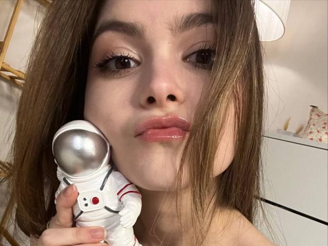 RonnaBarken with shiny lips, holding a white astronaut figure near her face, brown hair, and a white lamp in the background. Her hand is near her nose.
