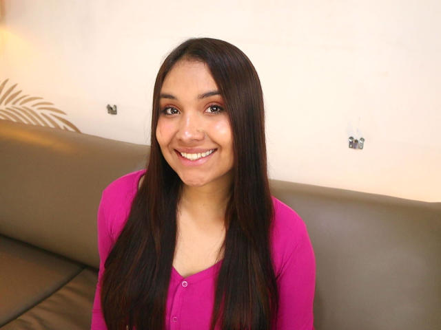 BelleLane has long dark brown hair, brown eyes, and a cute smile, wearing a pink long-sleeve shirt, sitting on a gray leather couch. Her tits are small.
