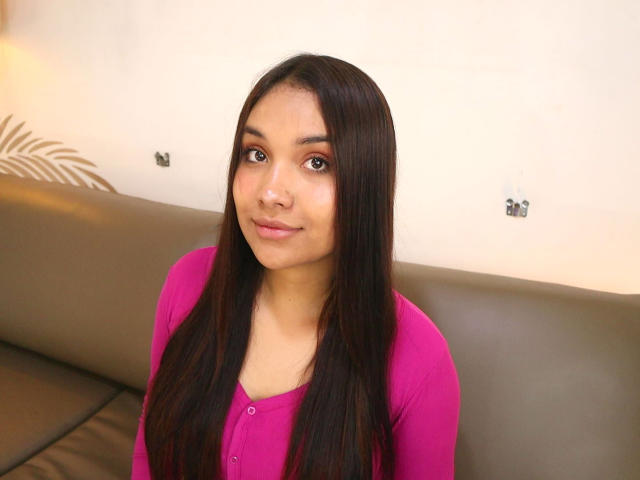 BelleLane has long, straight black hair, brown eyes, and a cute smile. She's wearing a pink cardigan and sitting on a gray leather couch.