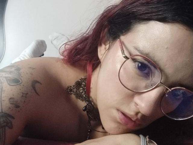 LaiaJonnes with red-dyed hair, glasses, and a black lace choker, lying down, showing tattoos on her shoulder, looking at the camera with a slightly parted lip.