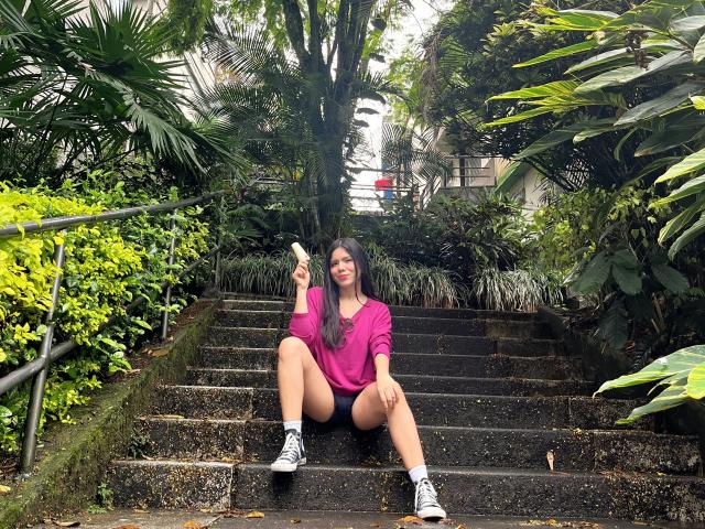 MaraCortes, sitting on wet stone stairs, wearing a pink sweatshirt, black shorts, and black sneakers, holding a popsicle, with a smile, legs spread, showing her ass.