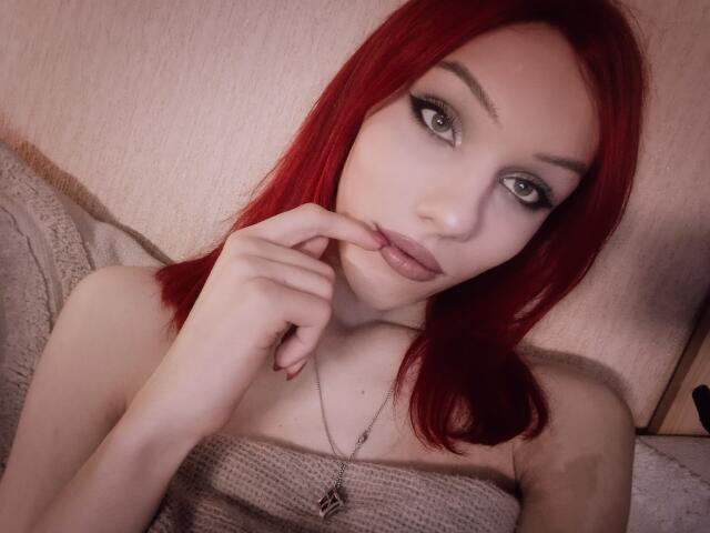 KiranaHayes with red hair, gray eyes, and pale skin, biting her finger, looking at the camera, wearing a strapless beige top, necklace with a small pendant.