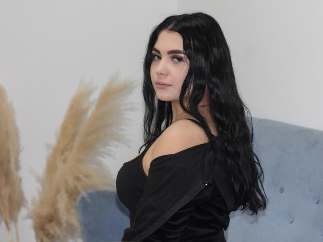 AdeleFelloy with long black hair, off-shoulder black top, looking back, showing her tits, sitting on a blue chair, puffy tan grass in the background.