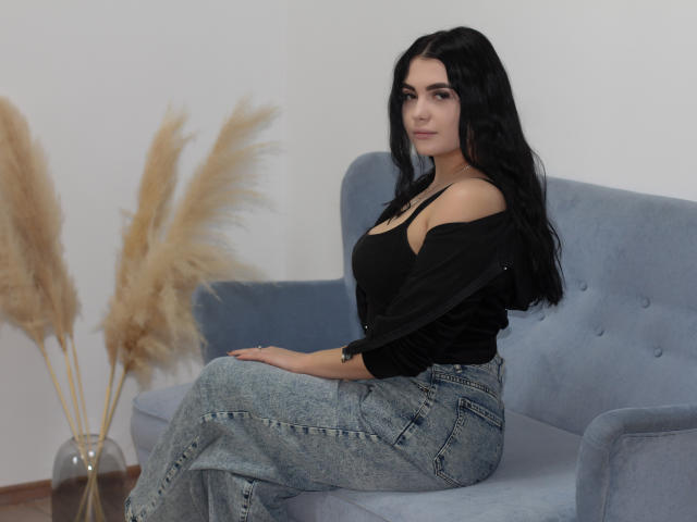 AdeleFelloy with long black hair, off-shoulder black top, and blue jeans, sitting on a blue sofa, puffy ass, slim build, and a relaxed, confident expression.