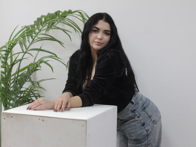 AdeleFelloy with long black hair, black velvet top, and high-waisted jeans leans on a white cube, green plant in background, looking at camera with a slight smile.