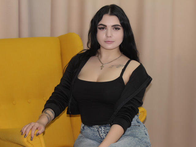 AdeleFelloy with long black hair, wearing a black top showing tits, denim jeans, and a yellow chair, has tattoos and a necklace, sitting with a relaxed, confident expression.