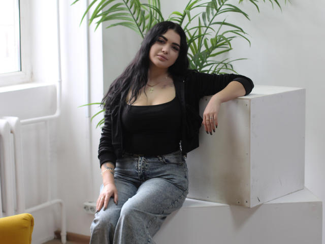 AdeleFelloy with long black hair, wearing a black top and jeans, sitting on a white cube, arms resting, looking slightly down, potted plant behind, yellow chair partially visible.