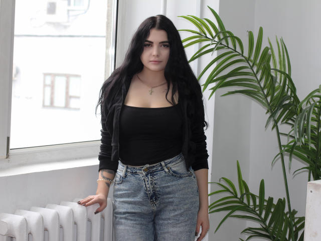 AdeleFelloy has long black hair, a black strapless top, and high-waisted blue jeans, standing by a window with a green plant, looking serious. She has small tits.