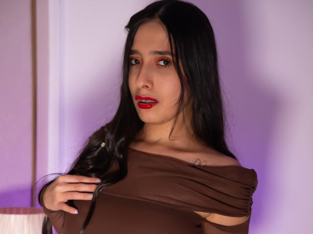 IvanaClark, avec de longs cheveux noirs, du rouge à lèvres, et un haut marron, tenant ses seins, petit tatouage sur le côté droit du cou, lumière violette douce en arrière-plan..