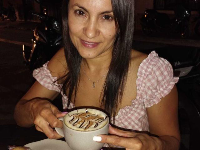 SolaraDubois with straight brown hair, pink checkered top, holding a foamy latte with chocolate leaf design, smiling at the camera, white nails, necklace, night setting.